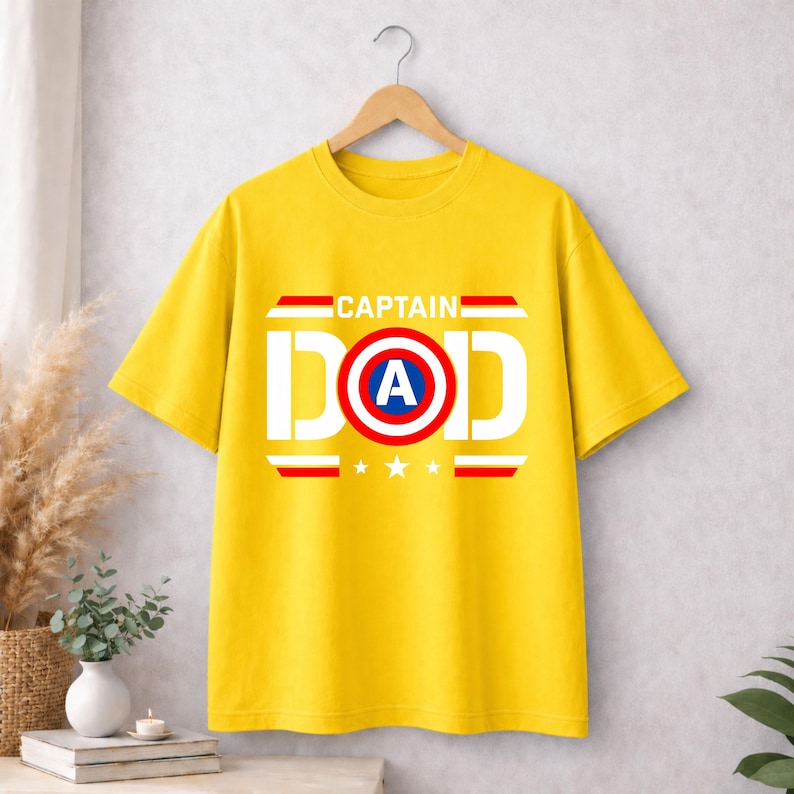 May include: A bright yellow short-sleeved t-shirt featuring the text CAPTAIN DAD in white block letters. The letter O is replaced by a circular red, white, and blue shield graphic with a white A in the centre, flanked by three white stars.