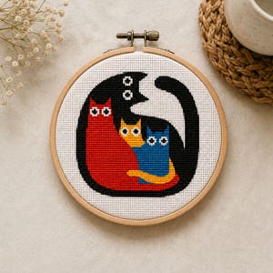May include: A cross-stitch embroidery hoop featuring a stylized family of cats. The design includes a large black cat embracing a red, yellow, and blue cat. The cats are set against a white background, framed by a wooden hoop.