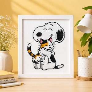 Può includere: Un ricamo a punto croce incorniciato raffigurante un cane bianco dei cartoni animati con orecchie nere e una macchia nera sulla schiena, che abbraccia affettuosamente un piccolo gatto calico con pelo arancione, nero e bianco. Design pixelato.