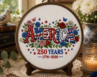 May include: A framed cross-stitch embroidery with the word "AMERICA" in red, white, and blue, surrounded by flowers and butterflies. The text "250 YEARS 1776-2026" is below. The embroidery is displayed on a wooden stand.