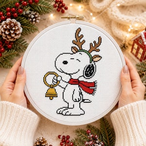 May include: A circular embroidery hoop containing a cross-stitch pattern of a white cartoon dog wearing brown reindeer antlers and a red scarf, holding a yellow bell. The design is stitched on white fabric with black outlines and details.
