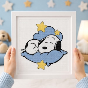 Peut inclure: Une broderie au point de croix encadrée représentant un beagle blanc aux oreilles noires dormant sur un oreiller bleu clair. Le motif comprend trois étoiles jaunes et un fond de nuage bleu clair, le tout dans un cadre carré blanc.