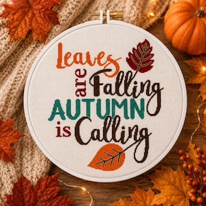 May include: A white fabric cross-stitch project in a round plastic hoop featuring the text "leaves are Falling AUTUMN is Calling" in orange, dark brown, and teal thread, accented with a dark red leaf and an orange leaf stitched on the fabric.