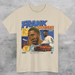 May include: Cream-colored t-shirt featuring a graphic design with the text "FRANK OCEAN" in blue and orange. The design includes illustrations of a person, a car, and the word "blond". The shirt has short sleeves and a crew neck.