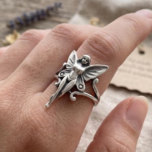May include: A silver-toned ring featuring a detailed fairy figure with outstretched wings. The fairy has a smooth, humanoid torso and defined wing patterns. The ring band is slender and curves around the finger, showcasing an oxidized finish.