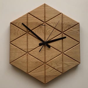 May include: A hexagonal wooden wall clock with a geometric design. The clock face is made of light-coloured wood, carved with a repeating triangular pattern. Black clock hands show the time. A modern, minimalist design.