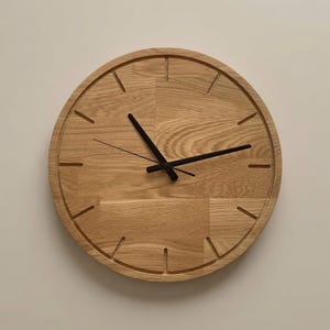 Op de afbeelding: Een ronde wandklok gemaakt van lichtbruin hout met een zichtbaar nerfpatroon. De wijzerplaat is voorzien van twaalf verzonken uurmarkeringen en drie dunne zwarte wijzers. Het ontwerp bestaat uit meerdere samengevoegde houtsegmenten.