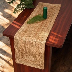 Peut inclure: Un chemin de table rectangulaire fabriqué en fibres de jute tressées de couleur beige naturel. Le chemin présente une texture tissée uniforme avec un détail de bordure. Une bougie pilier vert foncé et une branche de pin verte sont posées dessus.