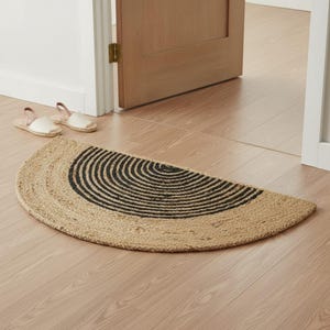 May include: A semi-circular woven jute rug featuring a natural tan fiber base with a series of concentric black circular lines radiating from the center. The rug is positioned on a light-colored wooden floor near a doorway with two sandals.