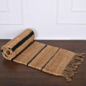 May include: A partially unrolled rectangular runner rug made of natural tan woven fibres featuring three distinct horizontal black stripes across the surface and finished with a fringe of loose fibre tassels along the short edge.