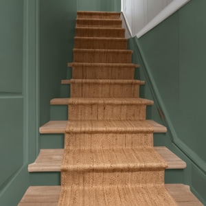 May include: A natural fibre stair runner with a tight, woven texture in a light tan colour. The runner is installed centrally over wooden stair treads, extending from the bottom step up to the top of the staircase, framed by sage green walls.