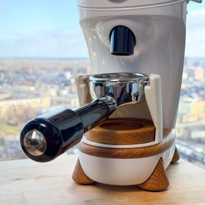 May include: A white coffee grinder with a black handle and a wooden base. The grinder has the word "NiCHE" on the top. The portafilter is inserted into the grinder. The grinder is on a wooden surface.