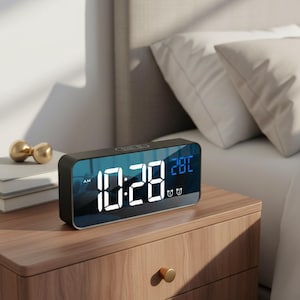 May include: A rectangular black digital alarm clock displaying the time 10:28 AM and the temperature 28°F. The clock has a mirrored surface and sits on a wooden nightstand. Two gold decorative spheres are visible in the background.