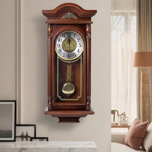 May include: A dark brown wooden wall clock with a pendulum. The clock face is white with black Roman numerals and gold hands. The clock has a decorative top and a shelf at the bottom. The clock is hanging on a light beige wall.