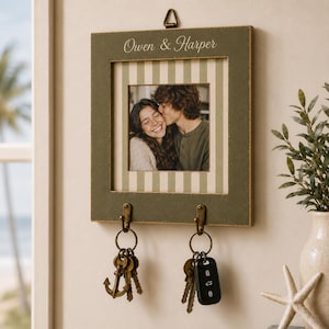 May include: A square olive green wall-mounted photo frame with a striped mat, featuring the text "Owen & Harper" at the top. The frame includes two bronze-colored metal hooks at the bottom, each holding a keyring with keys and an anchor charm.