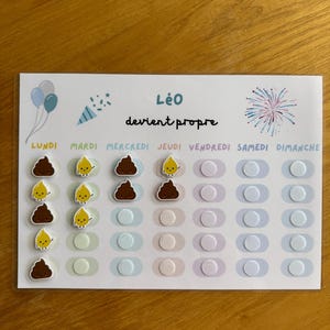 Peut inclure: Un tableau blanc avec le texte "Léo devient propre" et les jours de la semaine en français. Le tableau présente des illustrations de ballons, de confettis et de feux d'artifice. Il comprend des autocollants de caca souriant et de gouttelettes jaunes souriantes.
