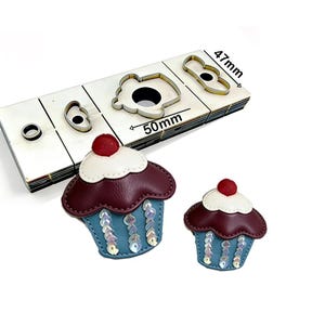 Può includere: Un set di fustelle in metallo a forma di cupcake, con dimensioni di 5 cm x 4,7 cm, accanto a due abbellimenti cupcake in pelle finiti. I cupcake presentano una base con paillettes blu, una parte superiore bordeaux, glassa bianca e una ciliegia rossa.