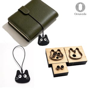 Puede incluir: Un cuaderno de cuero verde oliva con un dije negro con forma de gato en un cordón, un dije de gato negro separado en un cordón y dos bloques de madera con troqueles de metal con forma de cabeza de gato, con el logo de Ornaveda en la esquina.