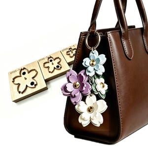 Puede incluir: Un bolso de mano de cuero marrón con un llavero floral. El llavero presenta dijes de flores moradas, blancas y azul claro con centros dorados. En el fondo se ven troqueles de madera con forma de flor.