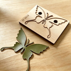 May include: A rectangular wooden cutting die with the text OT-0816 and a butterfly-shaped cutout, accompanied by a single olive green butterfly-shaped piece of material with a textured surface that matches the shape of the die cutout.