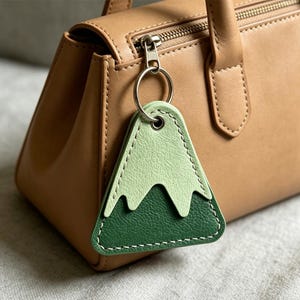 May include: A green and light green mountain-shaped leather keychain with white stitching, attached to a tan handbag with a silver zipper. The keychain is a triangular shape with a mountain design.