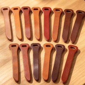 May include: A collection of fourteen flat, elongated leather pulls arranged in two rows. Each pull features a rounded top with a punched hole and a tapered, curved bottom edge, showcasing various shades of brown, tan, and reddish-brown leather.