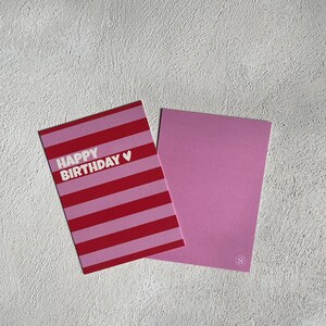 May include: A pink and red striped "Happy Birthday" greeting card with a white heart symbol. The card is next to a solid pink card with a small white "S" logo in the corner.