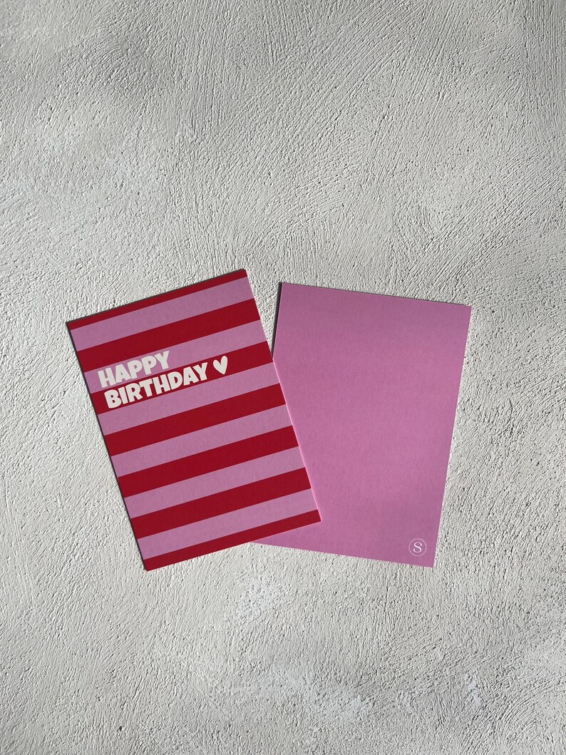 May include: A pink and red striped "Happy Birthday" greeting card with a white heart symbol. The card is next to a solid pink card with a small white "S" logo in the corner.