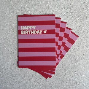 May include: A stack of birthday cards with a red and pink striped design. The cards have the words "HAPPY BIRTHDAY" in white, with a small heart symbol. The cards are fanned out on a white textured surface.
