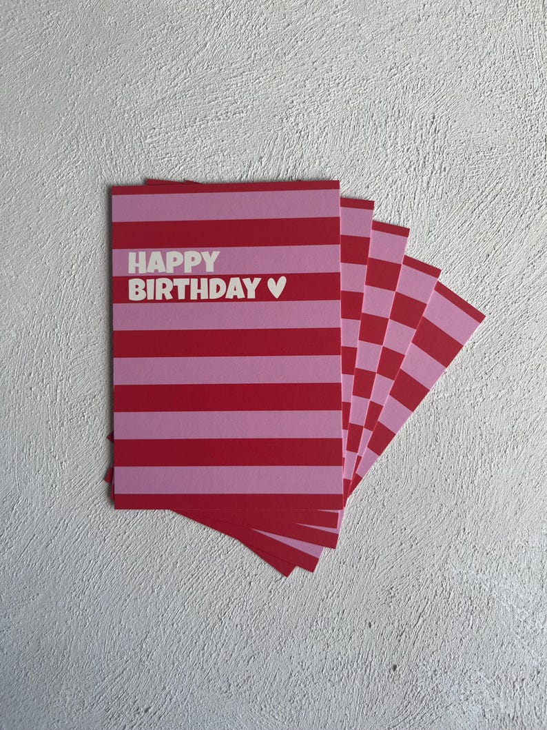 May include: A stack of birthday cards with a red and pink striped design. The cards have the words "HAPPY BIRTHDAY" in white, with a small heart symbol. The cards are fanned out on a white textured surface.