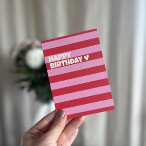May include: A birthday card with a red and pink striped pattern. The words "HAPPY BIRTHDAY" are printed in white, with a small heart symbol. The card is held in a hand.
