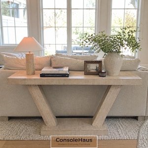May include: A light-colored wooden console table with a V-shaped base, topped with a lamp, books, a vase with greenery, and a framed picture. The table is set against a neutral-toned sofa and a large window.