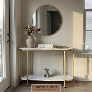 May include: A modern entryway console table with a white marble top and gold metal legs. The table has a lower shelf and is decorated with a vase of dried flowers, books, and a small tray. A round mirror hangs above.