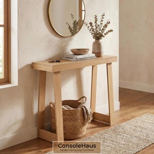 May include: A light wood console table with a rectangular top and angled legs. A woven basket sits below, and a round mirror hangs on the wall above. The table is decorated with books, a bowl, and a vase with dried flowers. The text "ConsoleHaus Modern Entryway Furniture" is at the bottom.