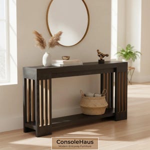 May include: A dark brown console table with vertical light brown accents. The table has a lower shelf and is decorated with a vase, books, a small sculpture, and a woven basket. A gold-framed mirror hangs above the table. The text "ConsoleHaus Modern Entryway Furniture" is at the bottom.
