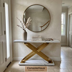 May include: A modern entryway table with a white marble top and gold-colored crossed legs. A round mirror hangs above the table, reflecting the room. A vase with dried plants and books are on the table. The text "ConsoleHaus Modern Entryway Furniture" is at the bottom.