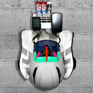 May include: A white wall-mounted cycling gear organiser holding a pair of white cycling shoes, a white helmet with a teal and pink visor, blue-tinted sunglasses, two black electronic cycling computers, and three cylindrical tubes of electrolyte tablets.