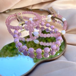 Könnte beinhalten: Eine Sammlung von Perlenarmbändern mit facettierten rosa und violetten Edelsteinperlen, glatten weißen ovalen Perlen und kleinen goldfarbenen metallischen Zwischenperlen. Jedes Armband enthält ein kleines Papieretikett mit handschriftlichem Text.