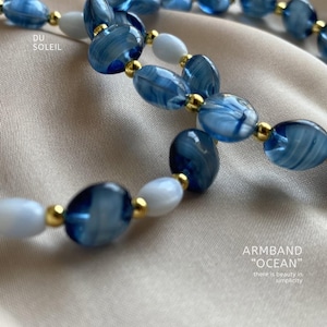 Könnte beinhalten: Ein Armband mit blauen und weißen ovalen Perlen, die durch goldfarbene Perlen unterbrochen werden. Das Armband ist mit "Ocean" beschriftet und der Text "Du Soleil" ist sichtbar. Der Hintergrund ist ein hellbeiger Stoff.