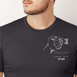 Könnte beinhalten: Dunkelgraues T-Shirt mit weißer Grafik auf der linken Brust. Die Grafik zeigt eine Faust, die die Hand eines Babys hält, mit Morsecode und dem Text "EST. 2020" darunter. Das T-Shirt hat kurze Ärmel und einen Rundhalsausschnitt.
