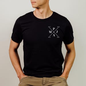 Könnte beinhalten: Schwarzes T-Shirt mit Rundhalsausschnitt und weißem Grafikdesign auf der Brust. Das Design zeigt sich kreuzende Linien mit den Buchstaben "P", "M", "J" und "A" sowie den Zahlen 2017, 2018, 2019 und 2022.