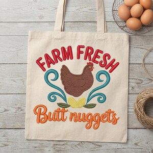 May include: A beige canvas tote bag featuring an embroidered design of a brown chicken, two yellow eggs, and teal swirls. The text reads Farm Fresh in red and Butt nuggets in orange. The bag has two top handles and a flat rectangular shape.