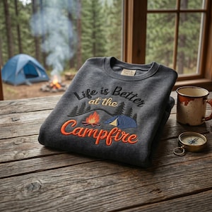 May include: A charcoal gray crewneck sweatshirt featuring embroidered text that reads Life is Better at the Campfire, accompanied by a graphic of a campfire, a blue tent, and black pine trees. The interior neck label reads Cabin & Trail Co.