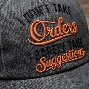 Op de afbeelding: Een grijze baseballcap met de geborduurde tekst "I DON'T TAKE Orders I BARELY TAKE Suggestions" in zwarte en oranje draad. De cap heeft een gebogen klep en metalen oogjes.