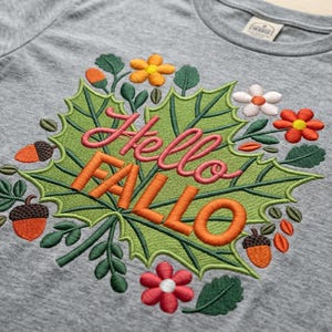 Könnte beinhalten: Ein graues T-Shirt mit einem gestickten Design. Das Design zeigt die Worte "Hello FALLO" in Rosa und Orange, umgeben von grünen Blättern, orangefarbenen Eicheln und bunten Blumen. Das Shirt ist aus einem weichen, bequemen Material.