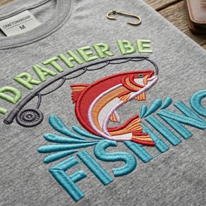 May include: Gray crew neck t-shirt with embroidered text and graphics. The design features the phrase "I'd Rather Be Fishing" in green and blue, with a leaping fish in red, orange, and white. A fishing rod is also included in the design.
