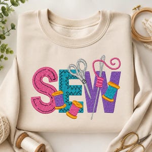May include: A beige crewneck sweatshirt featuring an embroidered design on the front. The word SEW is displayed in large, colorful letters. The S is pink, the E is teal, and the W is purple. A silver pair of scissors and a needle with pink thread are integrated into the letters, accompanied by small colorful spools of thread.