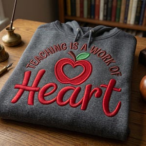 May include: A charcoal gray hooded sweatshirt featuring red embroidered text that reads Teaching is a work of and a large red apple with a heart-shaped center and a green leaf. The word Heart is embroidered in large red script below the apple.