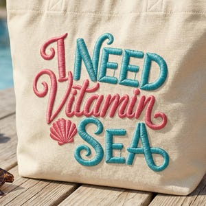 Peut inclure: Un sac fourre-tout beige avec la phrase brodée "I NEED Vitamin SEA" en rose et turquoise. Le sac est posé sur une terrasse en bois à côté de lunettes de soleil, d'un coquillage, d'une serviette corail roulée et d'un verre de thé glacé.