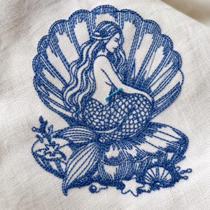 May include: A blue thread embroidery design on light-colored fabric featuring a mermaid sitting inside a large open seashell. The mermaid has long flowing hair and a scaled tail, surrounded by small starfish and smaller shells at the base.
